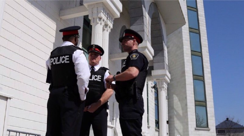 Three uniformed police officers stand conversing outside a white building with ornate columns under a clear blue sky.