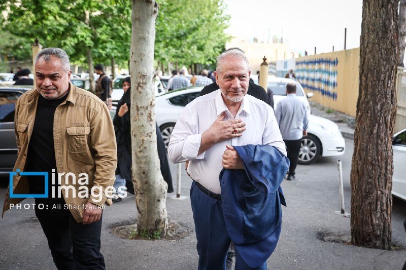 A bearded man in a white shirt walks on a tree-lined sidewalk holding a blue jacket, accompanied by another man, with parked cars and pedestrians visible in the background.