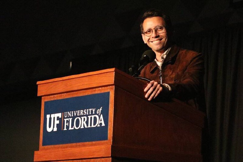 A smiling man in glasses stands at a wooden University of Florida podium, speaking into a microphone against a dark curtain backdrop.