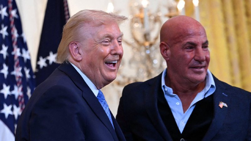 Two men in suits smile together in an ornate room featuring American flags, a chandelier, and gold curtains.