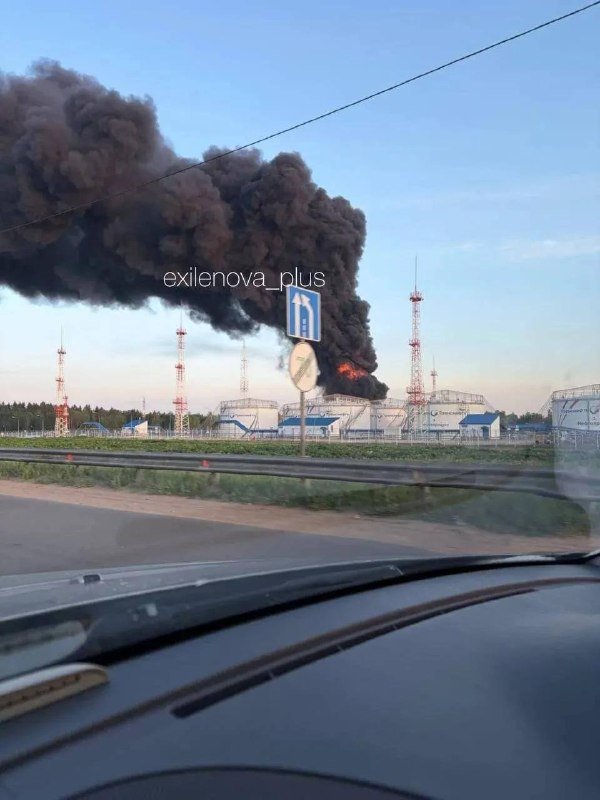 Thick black smoke billows from a fire near large white storage tanks and communication towers, viewed from inside a vehicle along a roadside.