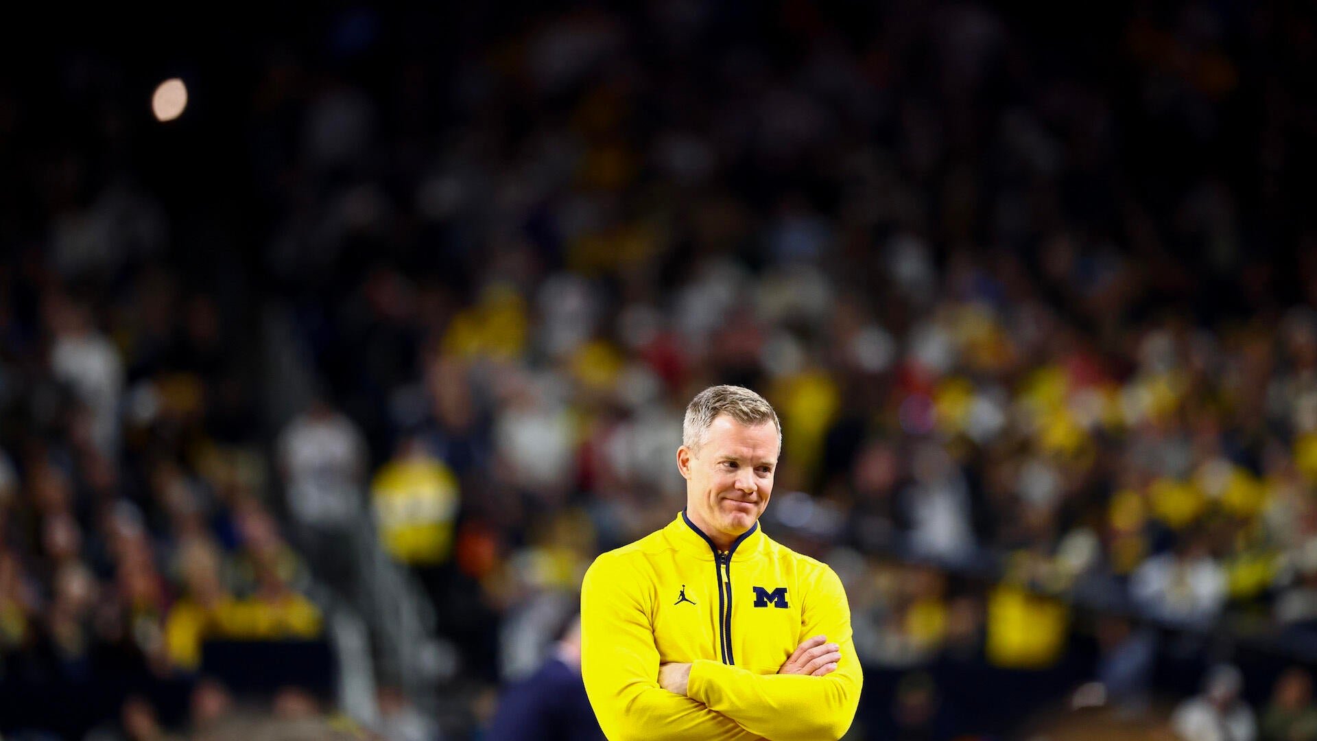 A man in a yellow quarter-zip pullover with an "M" logo stands with crossed arms before a blurred crowd in a stadium.