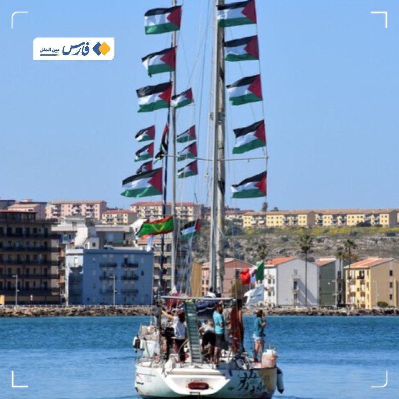 A sailboat adorned with numerous Palestinian flags sails on blue water, with people aboard and a coastal town visible in the background.