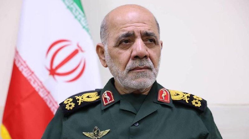 A gray-bearded man in a green military uniform with gold and red insignia stands in front of an Iranian flag.