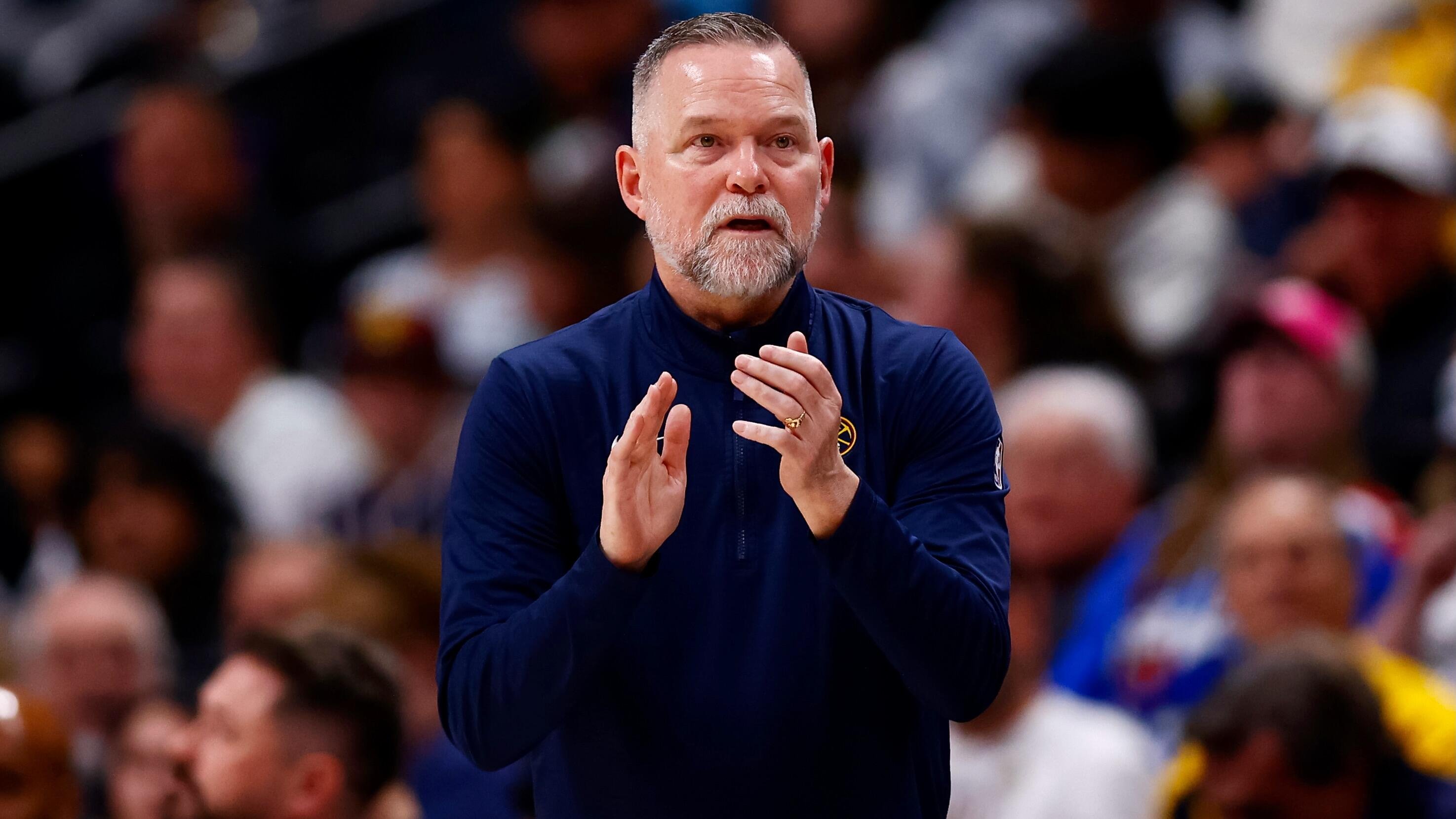 A gray-bearded man in a navy blue tracksuit claps his hands while standing in front of a blurred crowd.