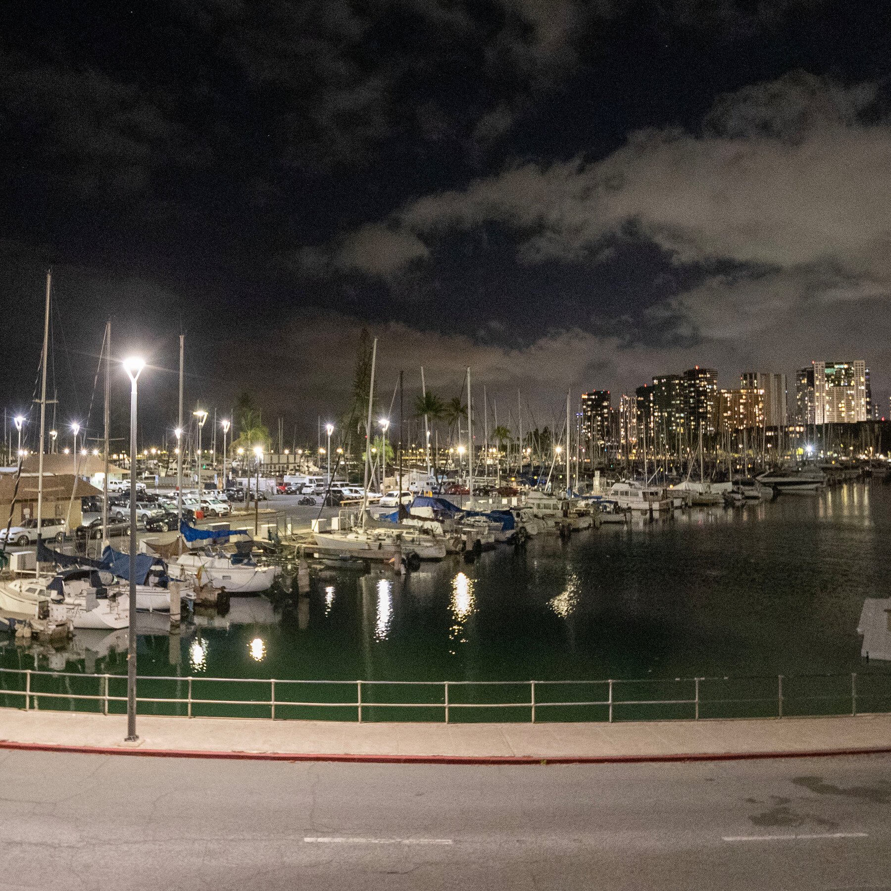 Sailboats are docked at a marina at night, with illuminated high-rise buildings and parked cars visible in the background under cloudy skies.