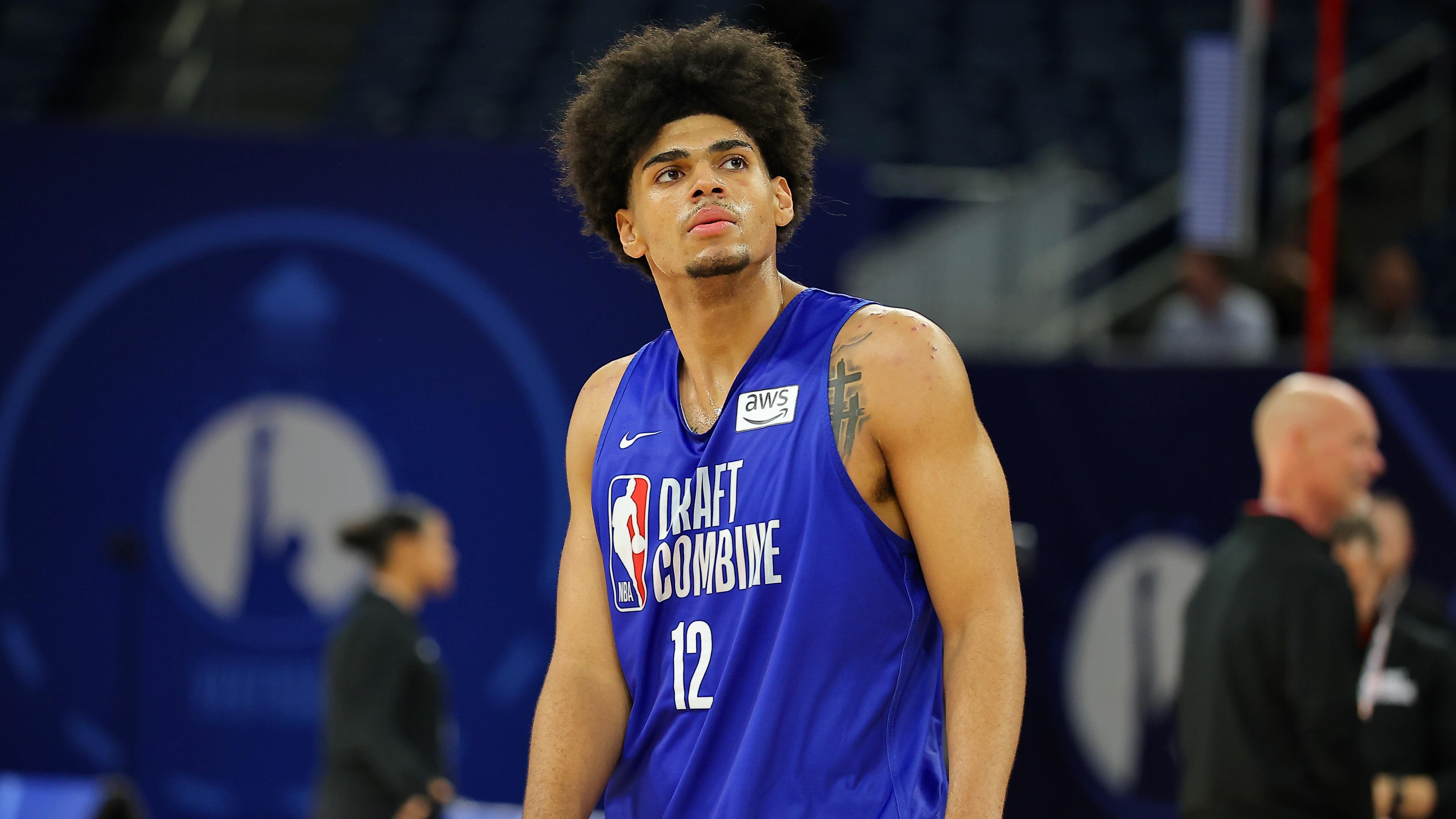 A young basketball player wearing a blue "Draft Combine" jersey numbered 12 stands on a court, looking upward during an NBA Draft Combine event.
