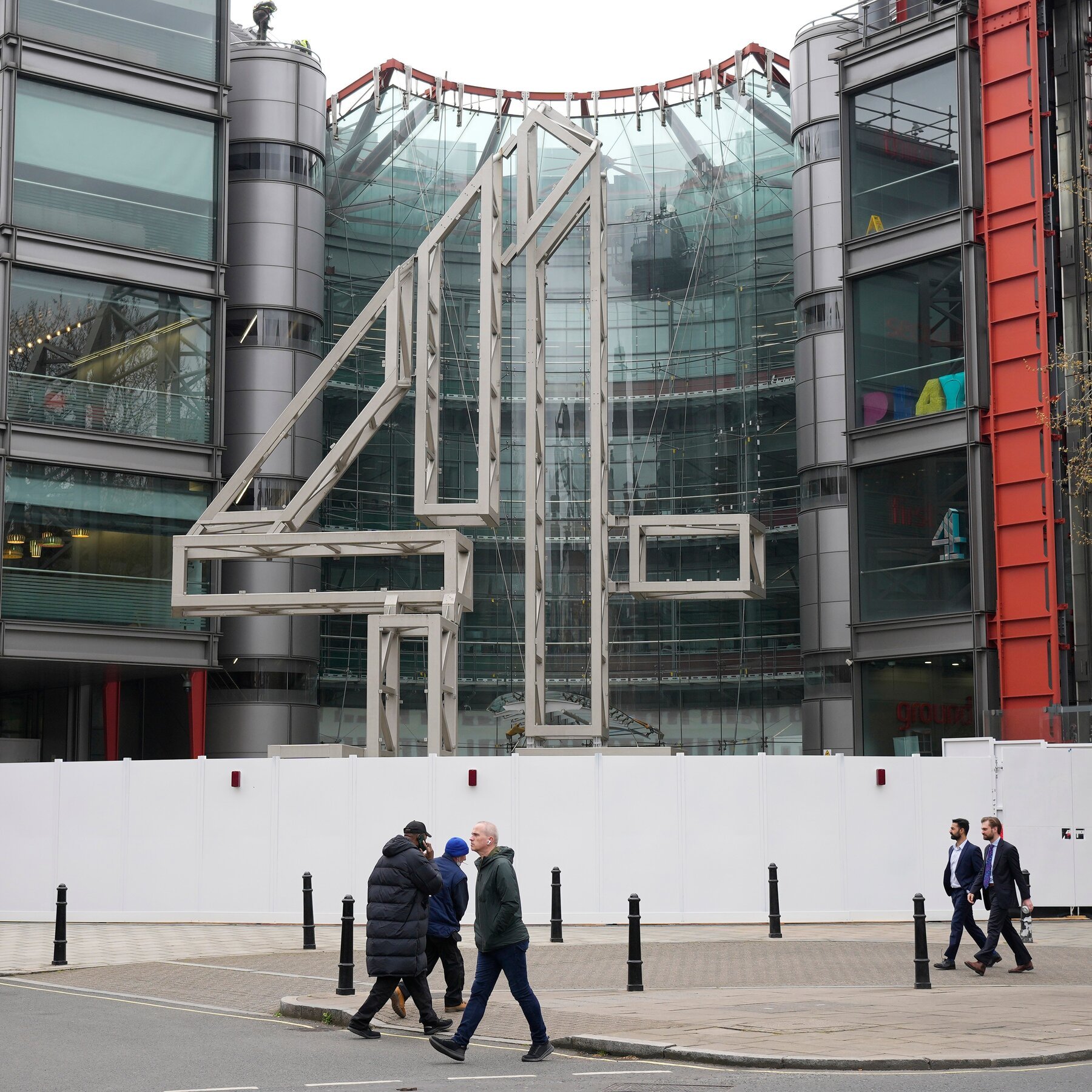 Pedestrians walk past a white construction barrier in front of a modern glass-and-steel building featuring a large metal geometric sculpture.