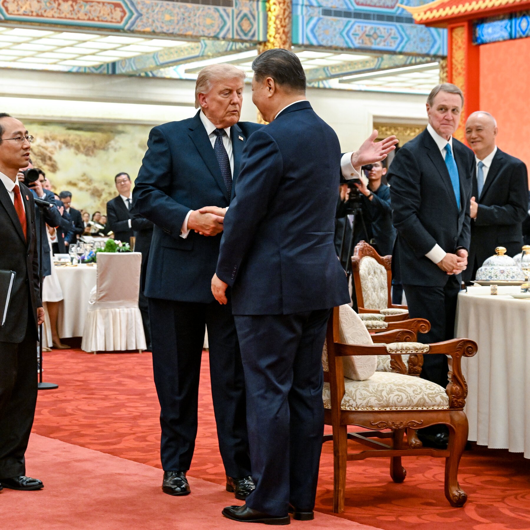 Two men in dark suits shake hands in an ornate room with decorated ceilings, red carpet, and round tables, while other formally dressed individuals stand nearby.