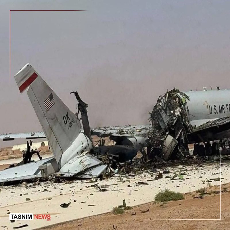 A destroyed U.S. Air Force aircraft lies in pieces on a debris-strewn tarmac, with its torn tail fin showing an American flag and scattered wreckage extending across the airfield.