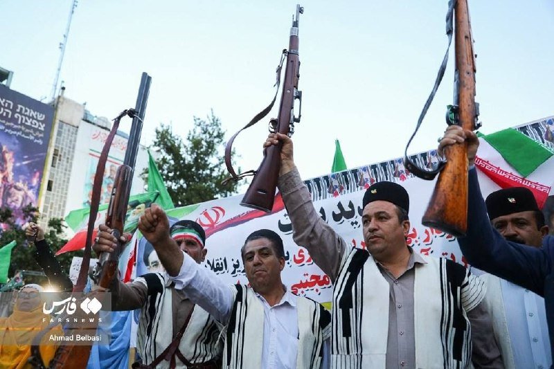 Men in traditional vests and caps raise rifles overhead during a rally with Persian-scripted banners and Iranian flags visible in the background.