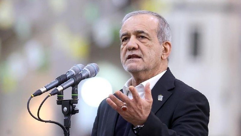 A gray-haired man in a dark blazer speaks at a microphone stand, gesturing with his hand, in an outdoor setting with a blurred background.