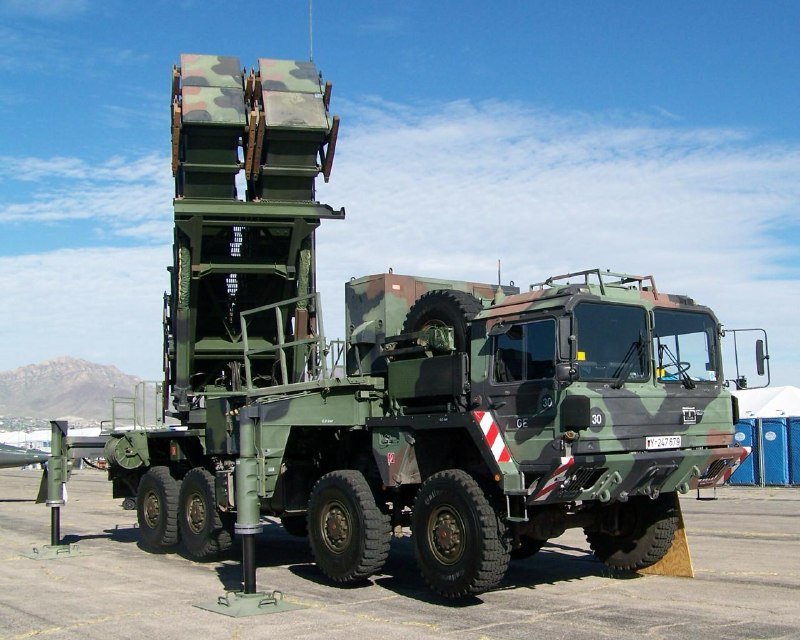 A camouflage-painted military truck carrying an elevated, vertical launcher system is parked on a paved lot with mountains visible in the background under a partly cloudy sky.