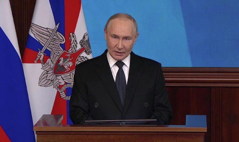 A gray-haired man in a black suit and tie speaks at a podium, with a Russian flag and emblem visible in the background.