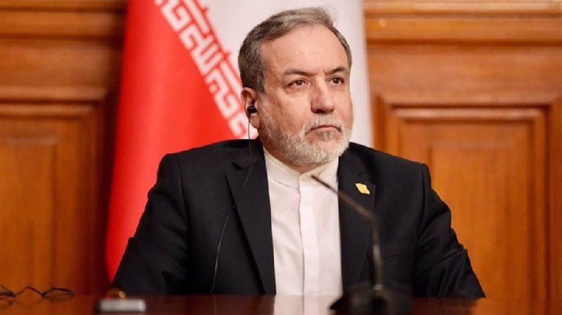 A gray-bearded man in a dark suit and white shirt sits at a table with a microphone, with a red and white flag visible behind him.