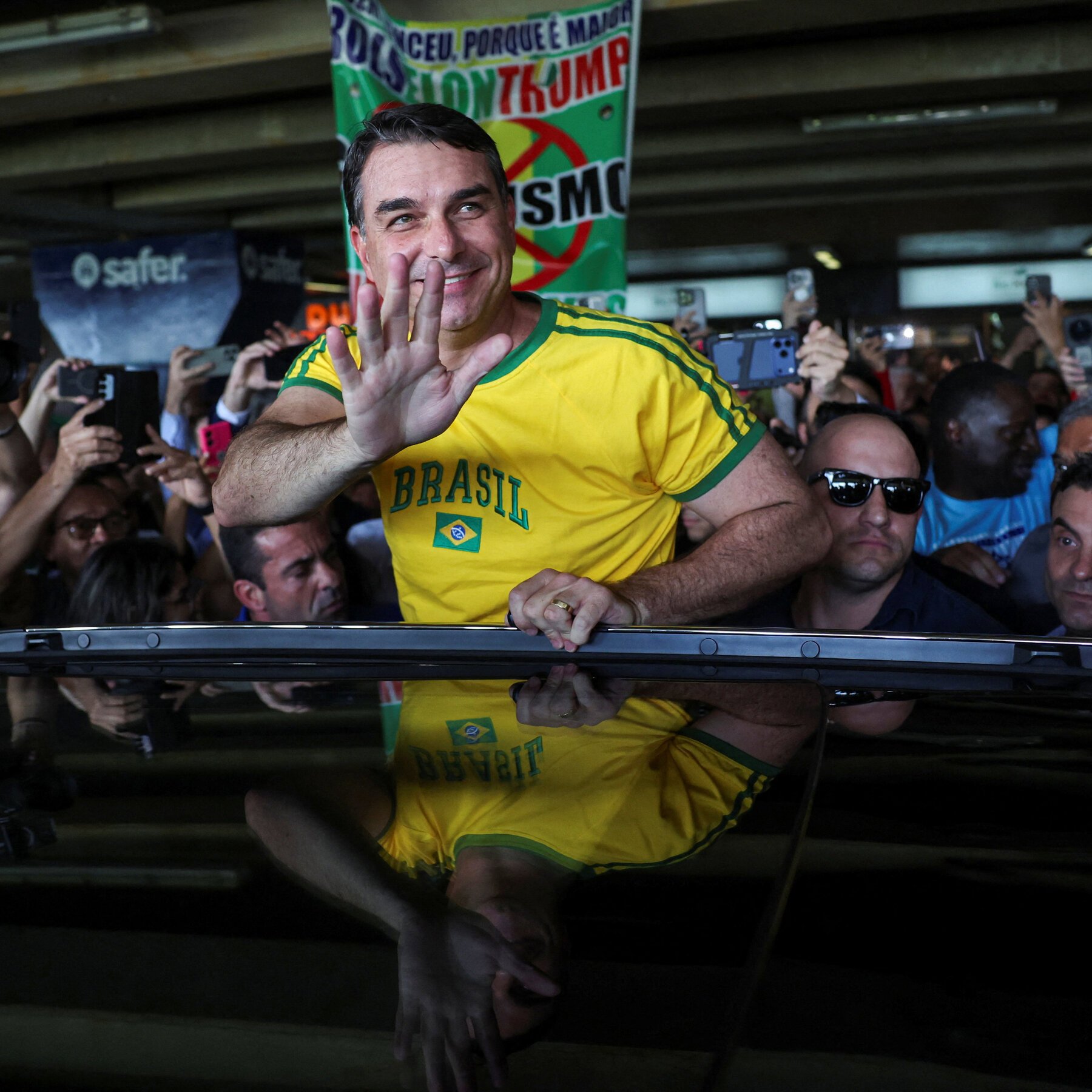A smiling man in a yellow Brazil jersey waves from a sunroof, surrounded by a crowd taking photos with phones.