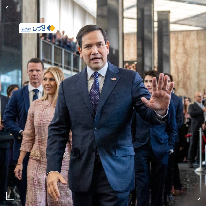 A man in a dark suit waves toward the camera while walking through a modern building, followed by a woman in a pink tweed outfit and several other people in formal attire.