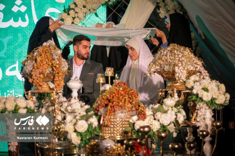 A bride and groom sit together under a white cloth held overhead at a ceremony, surrounded by elaborate floral arrangements and decorated tables.