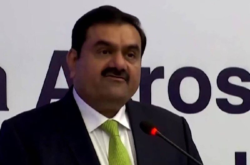 A man in a dark suit and green tie speaks into a microphone at a podium, with large letters partially visible behind him.