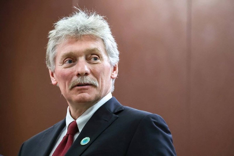 An older man with gray hair and a mustache, wearing a dark suit, white shirt, red tie, and a green lapel pin, looks off to the side against a brown background.
