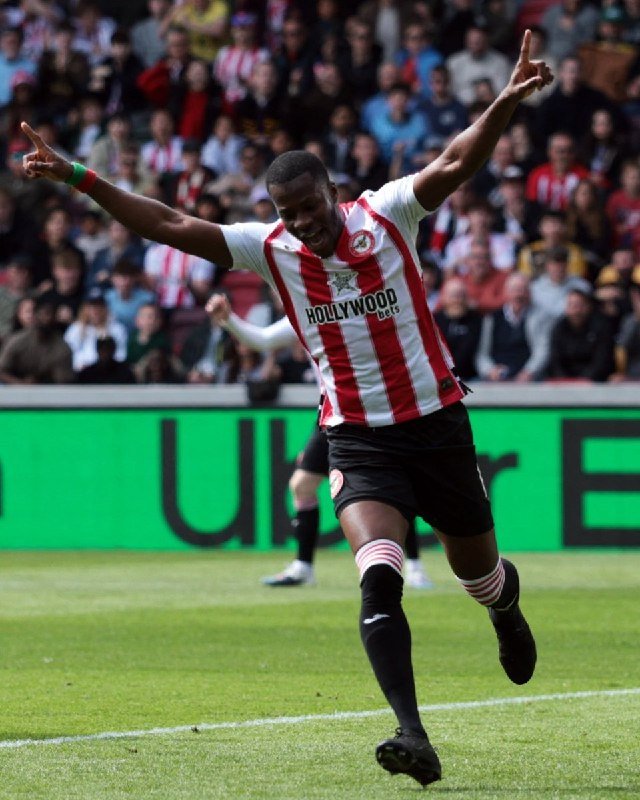 A soccer player in a red and white striped jersey with "Hollywoodbets" sponsorship runs with arms raised and fingers pointed upward in celebration before a crowded stadium.