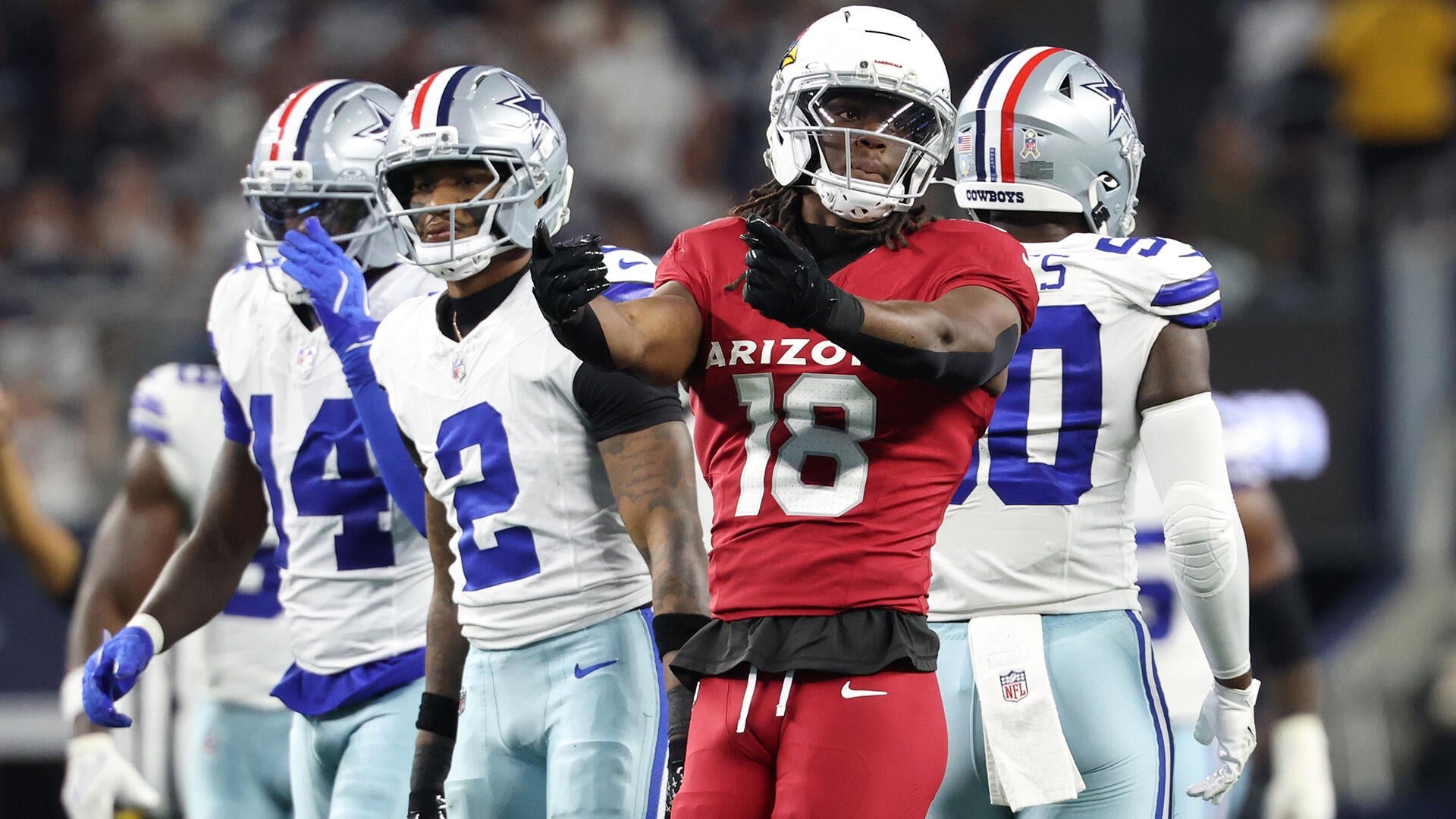 An Arizona Cardinals player wearing jersey number 18 in a red uniform gestures with both hands while standing near three Dallas Cowboys players in white uniforms on a football field.