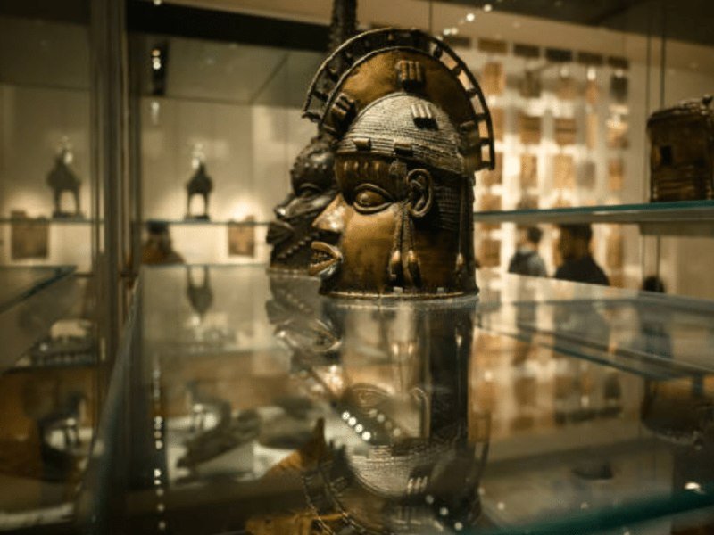 A bronze sculpted head with a halo-like crown is displayed inside a glass museum case, with other artifacts visible on shelves in the background.
