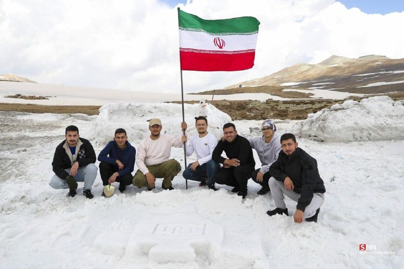 Seven men crouch in snow on a mountainous terrain, with one holding an Iranian flag, gathered around a heart shape drawn in the snow.