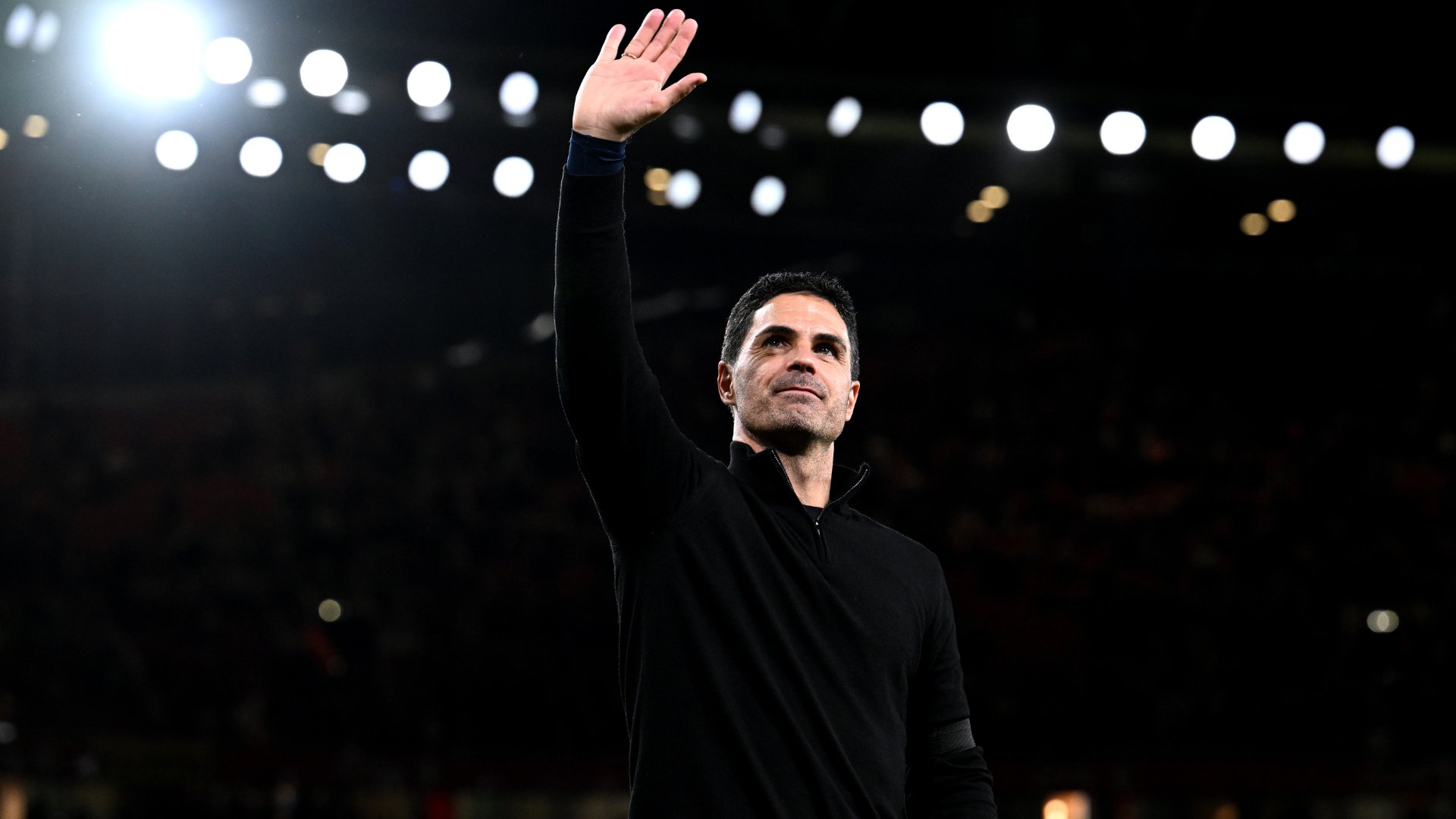 A man in a black long-sleeved top raises his right hand to wave toward a crowd, with stadium lights visible overhead against a dark background.