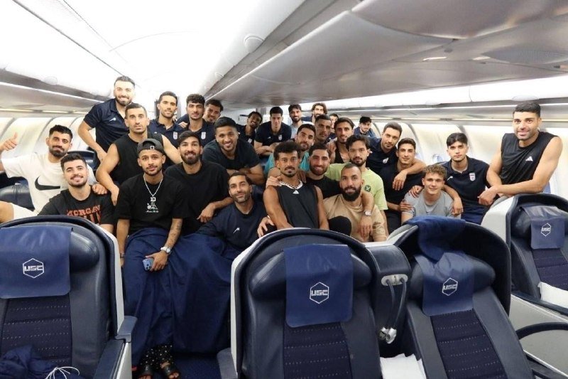 A large group of men, many wearing matching dark athletic attire, pose together for a photo inside a commercial airplane cabin with "USG" branded headrests.