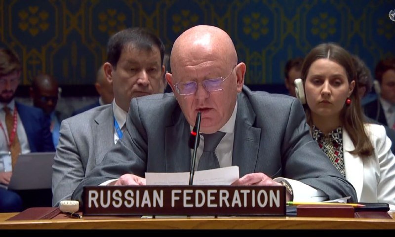 A bald man in a gray suit speaks into a microphone behind a "RUSSIAN FEDERATION" placard at a formal meeting, with other attendees seated nearby.