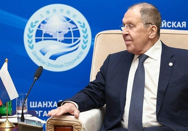 A bespectacled man in a dark suit and patterned tie sits in a beige chair before a microphone, with a Shanghai Cooperation Organization emblem and Russian flag visible in the background.