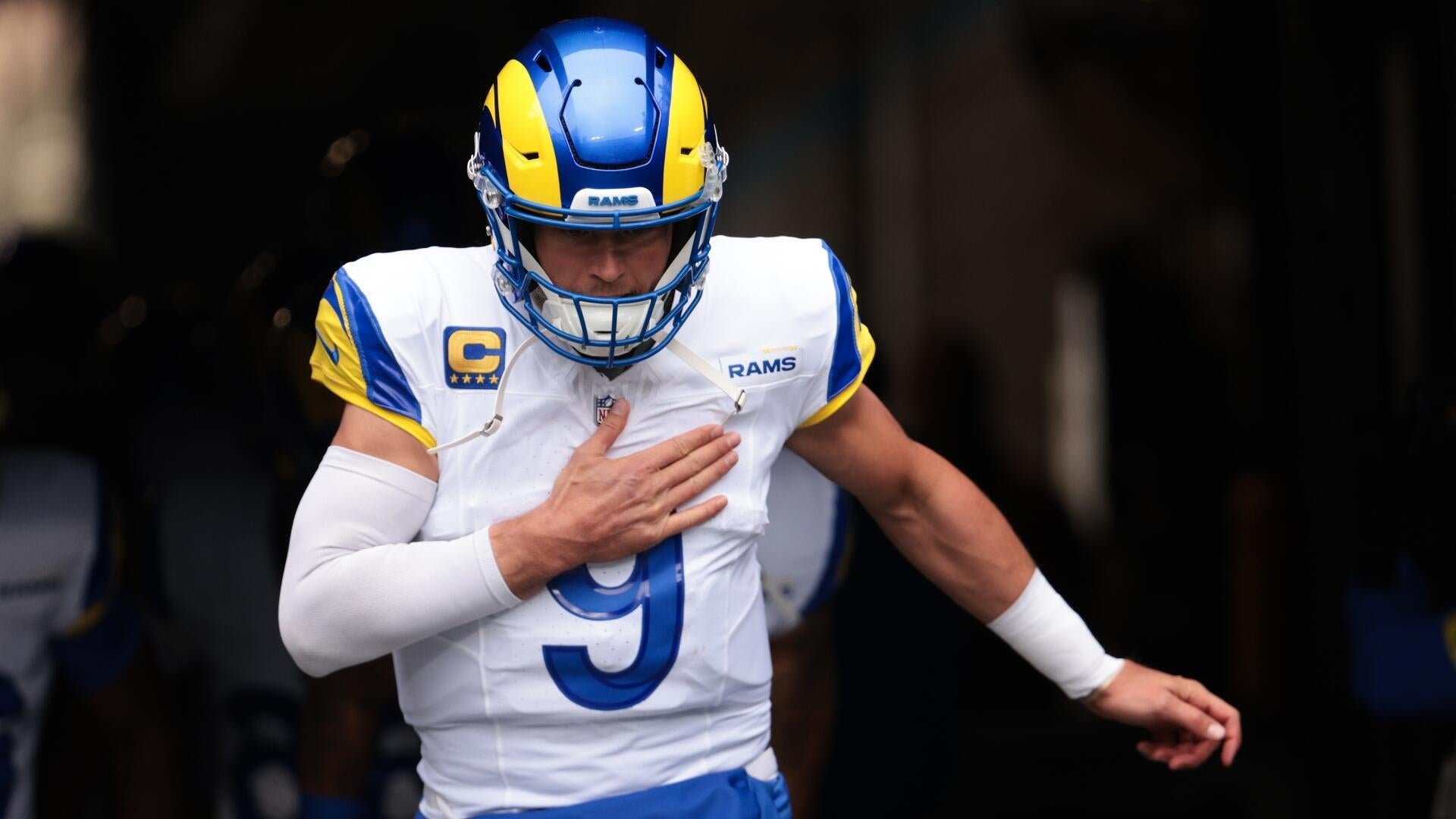 A football player in a white and blue Rams uniform and helmet runs forward with his hand on his chest.