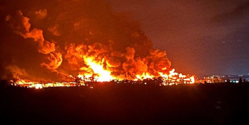 Massive orange flames and thick smoke billow into the night sky above an industrial area in the distance.