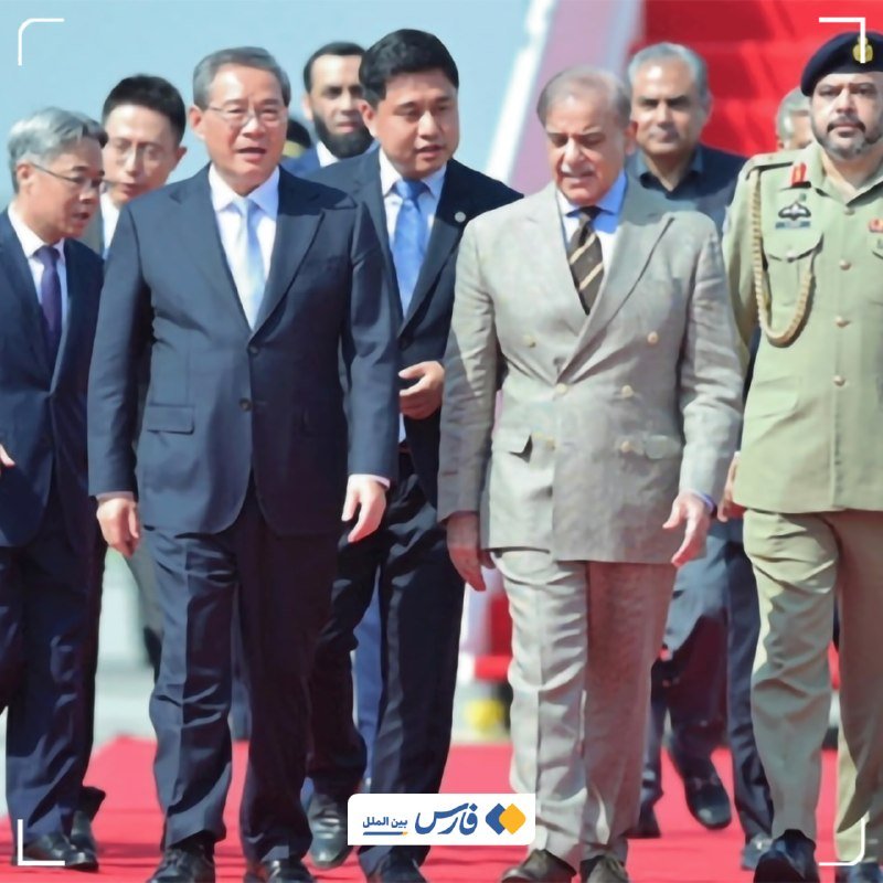 A group of men in business suits and a uniformed officer walk together along a red carpet during an outdoor official ceremony.