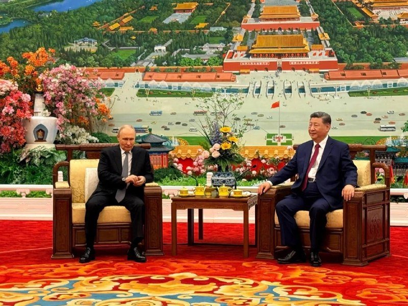 Two men in dark suits sit in ornate wooden chairs across a small table, with a large mural depicting a traditional palace complex behind them.