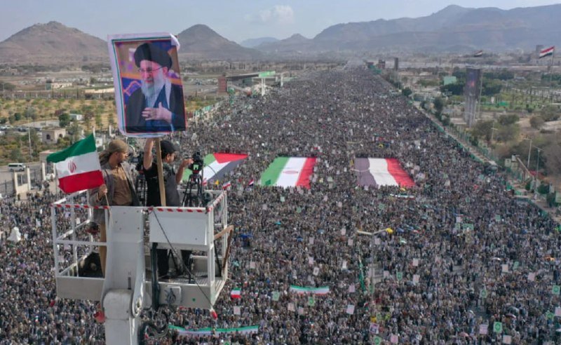 A massive crowd fills a wide road flanked by mountains, holding large Iranian flags and banners, while a worker in an elevated cherry picker films alongside a large framed portrait of a bearded cleric.