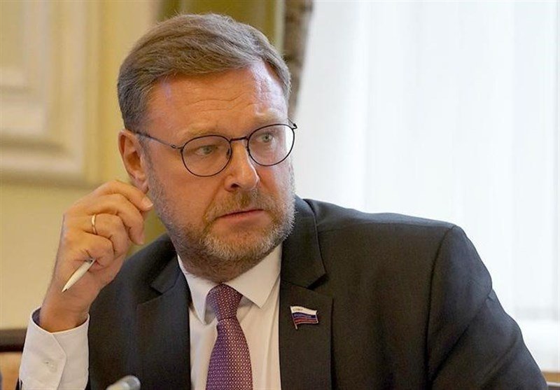 A bearded man wearing glasses, a dark suit, and a lapel pin listens while holding a pen near his face in a formal indoor setting.