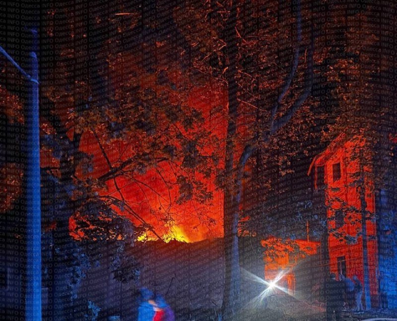 A large fire glows bright orange through silhouetted trees behind a stone wall at night, illuminating a building and a streetlamp, with a few people standing nearby.