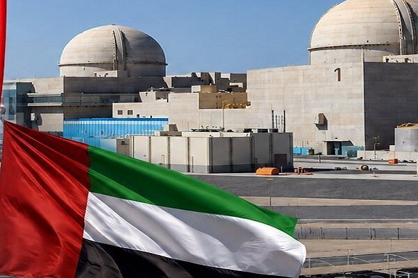 The United Arab Emirates flag waves in the foreground with a large nuclear power plant facility, featuring two domed reactor containment buildings, visible in the background under a clear sky.