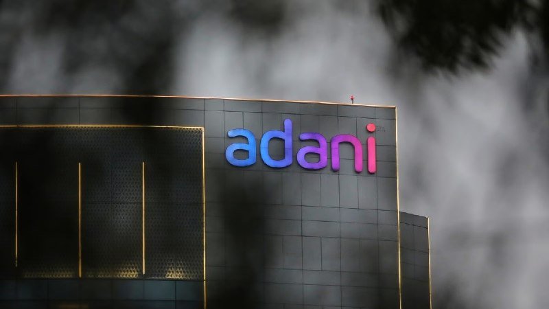 A building facade displays an illuminated "adani" logo in blue-to-pink gradient lettering, partially obscured by out-of-focus dark tree branches against a cloudy sky.