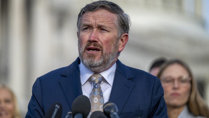 A bearded man in a navy suit and patterned tie speaks at an outdoor podium with microphones, while others stand behind him against a stone building.