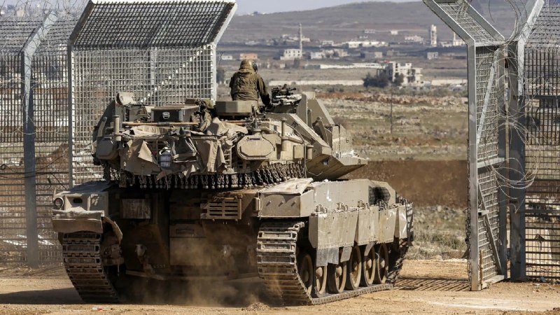 A soldier sits atop a tank as it passes through an open metal gate topped with barbed wire, with a town and hills visible in the distance.