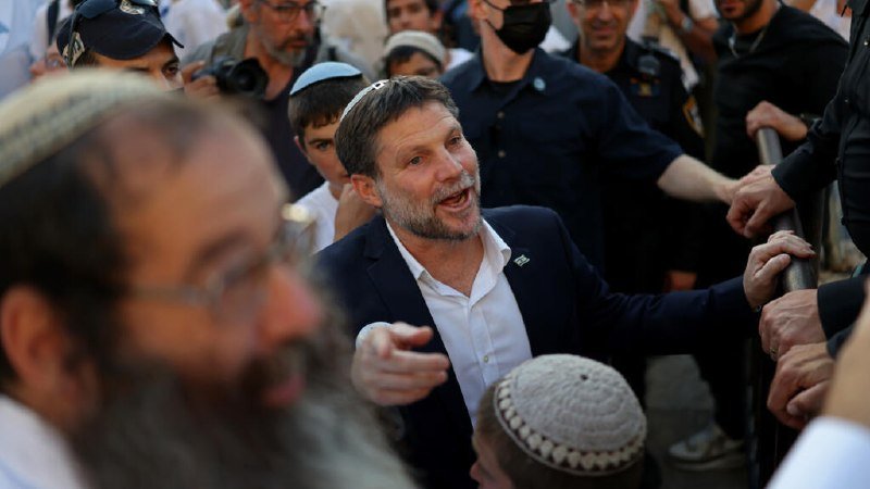A man wearing a kippah gestures while speaking to a crowd, with another bearded man in a kippah in the foreground.