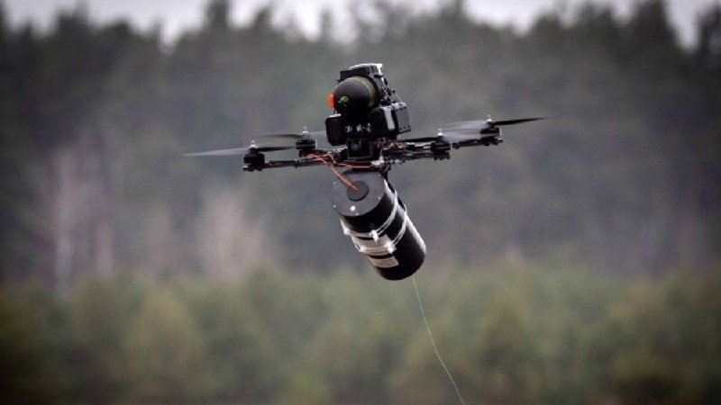 A black quadcopter drone carrying a cylindrical payload hovers mid-air against a blurred background of trees.