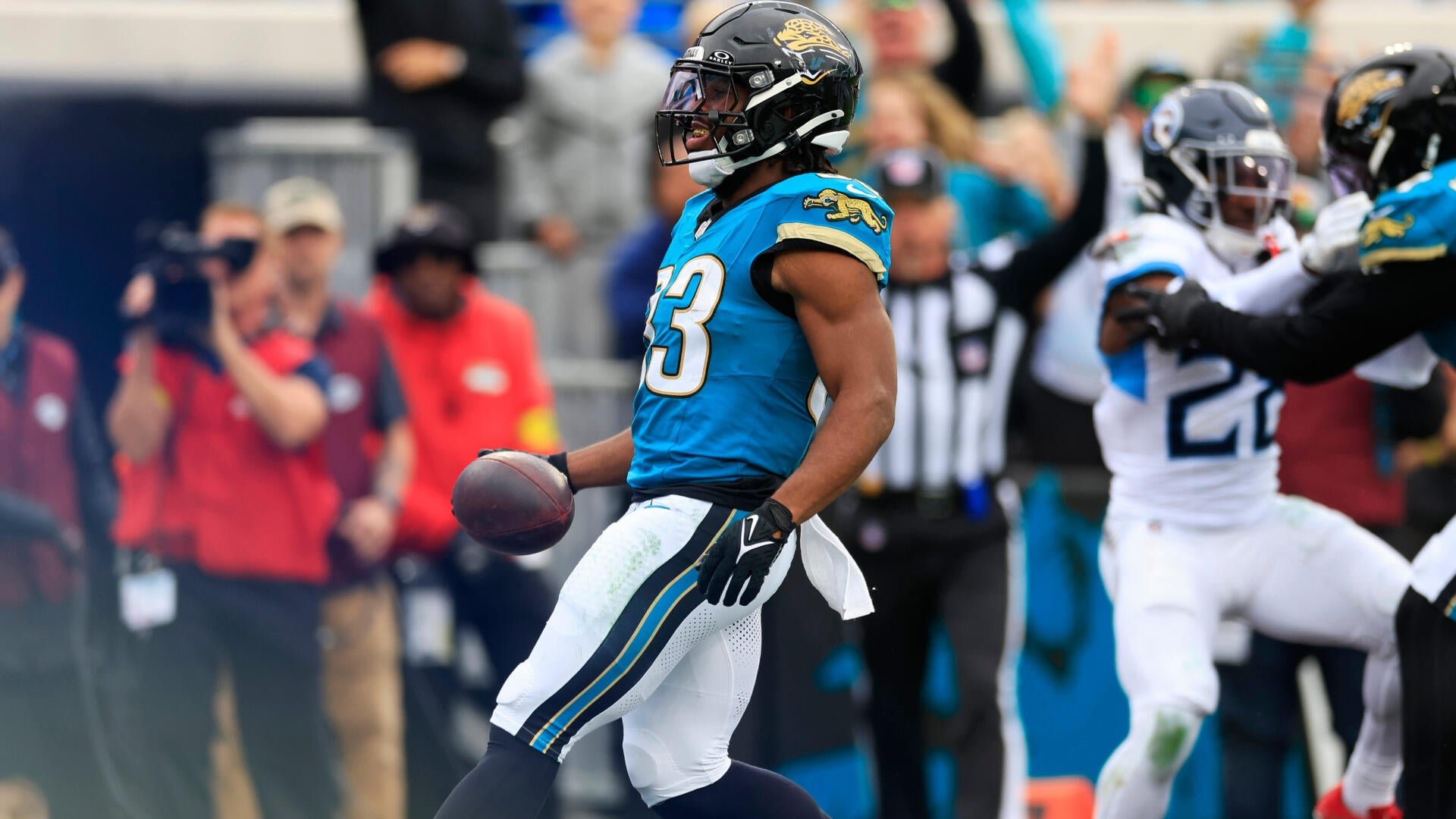 A Jacksonville Jaguars player in a teal jersey celebrates while holding a football, with a referee and opposing players visible in the background.