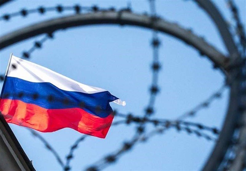 A white, blue, and red flag waves in the foreground, viewed through out-of-focus coils of barbed wire against a pale blue sky.