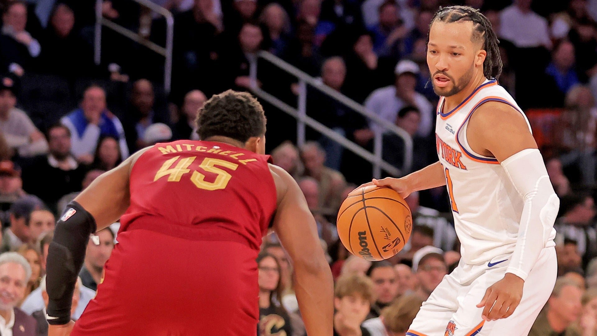 A Knicks player in a white jersey dribbles a Wilson basketball while facing a Cavaliers defender wearing a red "MITCHELL 45" jersey during a game in front of a crowd.