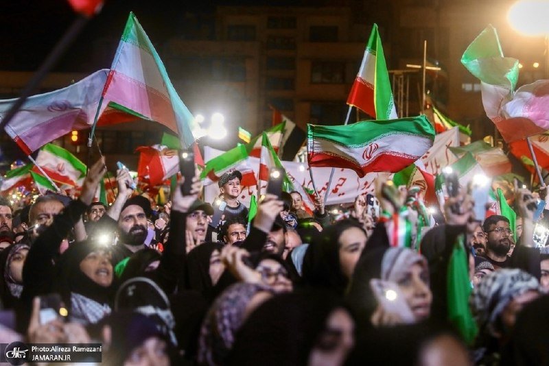 A nighttime crowd waves green, white, and red Iranian flags and holds up phone flashlights, with a child seated on someone's shoulders amid the gathering.