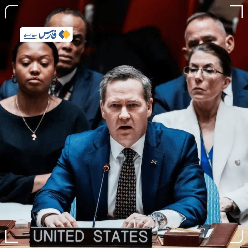 A man in a blue suit and patterned tie sits at a desk labeled "UNITED STATES," speaking into a microphone while several people sit behind him.